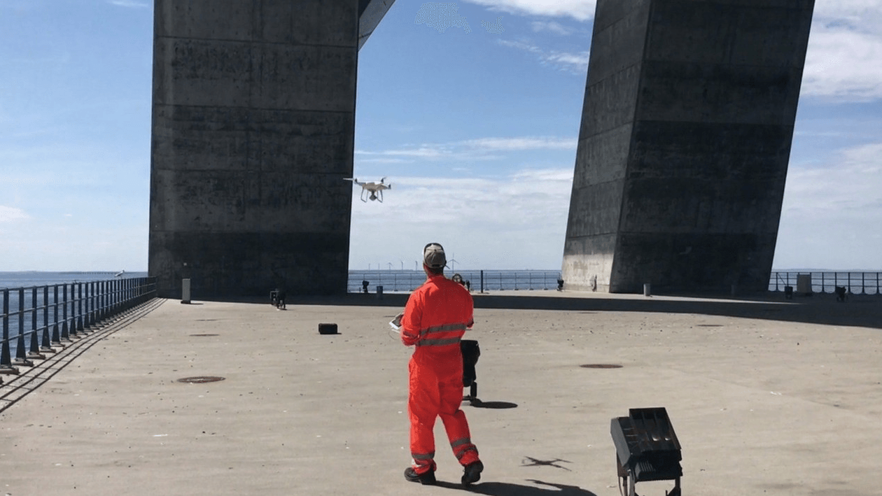 Photograph of a man in an orange work suit operating a drone under the pillars of a bridge