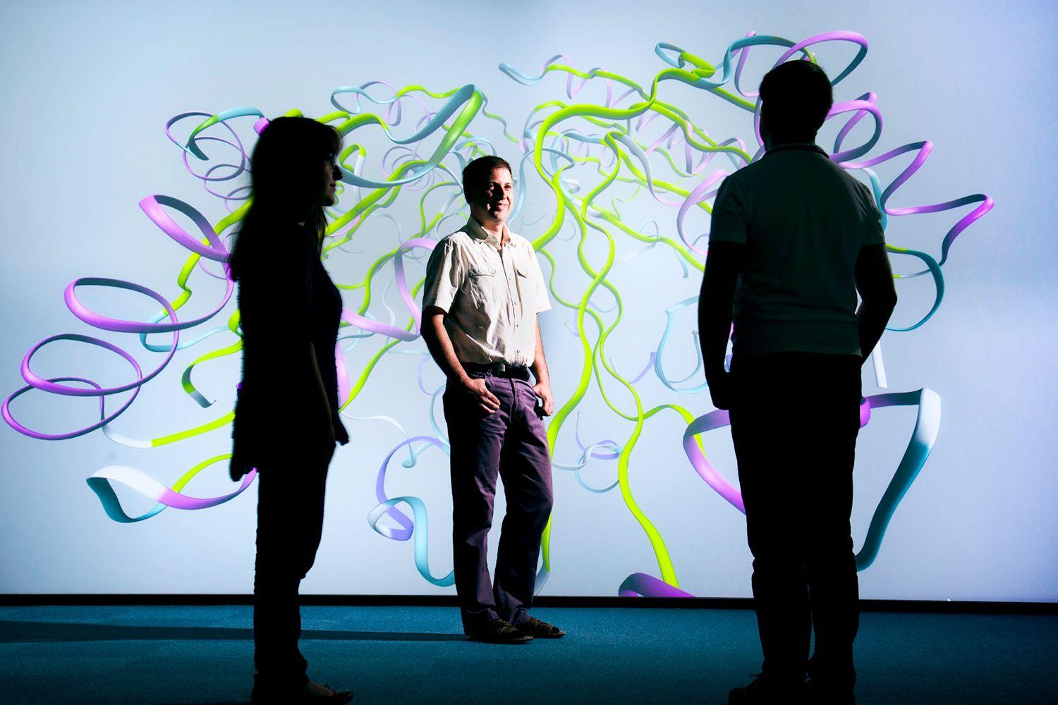 Photograph of the visualisation suite at the Hartree National Centre for Digital Innovation with three people against a backdrop of full screen wall with a ribbon diagram of a molecular structure